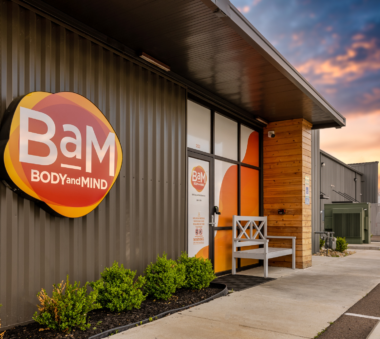 Exterior view of a modern building with a sign that reads 'BAM Body and Mind' and a bench near the entrance at sunset