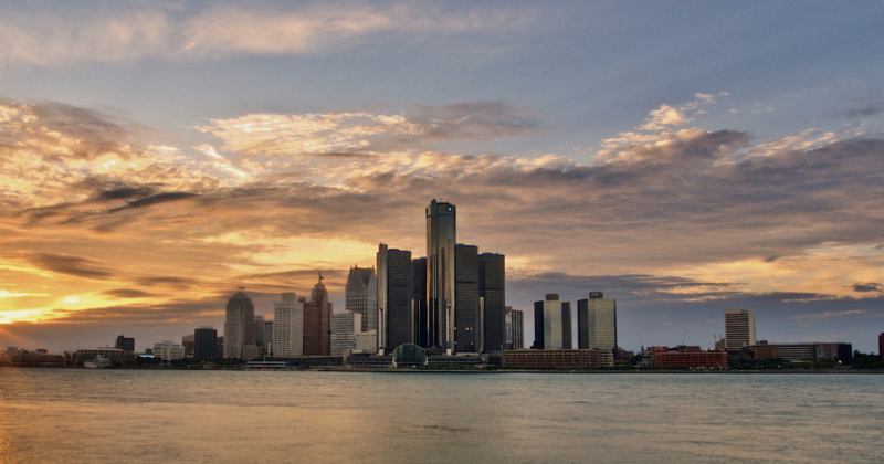 Detroit skyline at sunset with tall buildings reflecting light across the river.