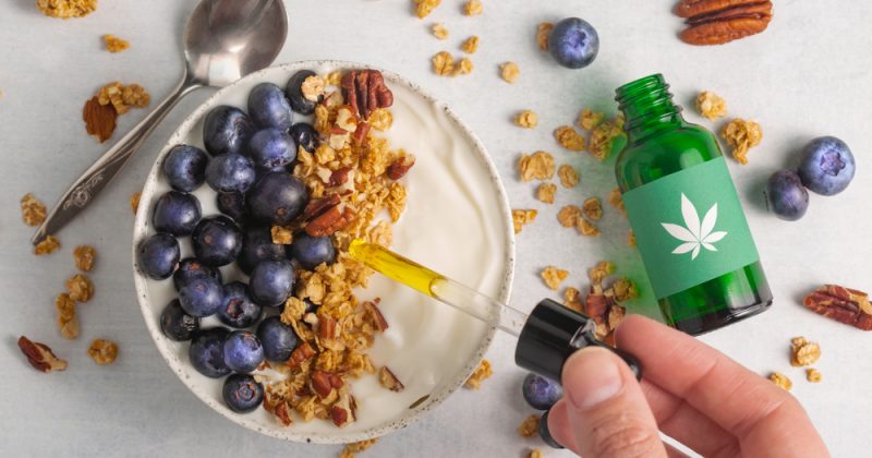 A hand adds CBD oil to a yogurt bowl topped with blueberries, granola, and pecans