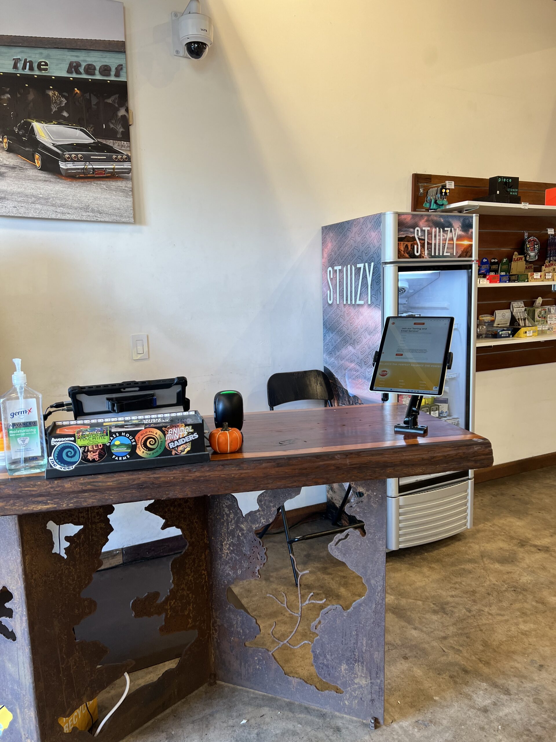 A wooden counter with hand sanitizer, a small pumpkin, and a touchscreen device in front of a branded refrigerator and shelves