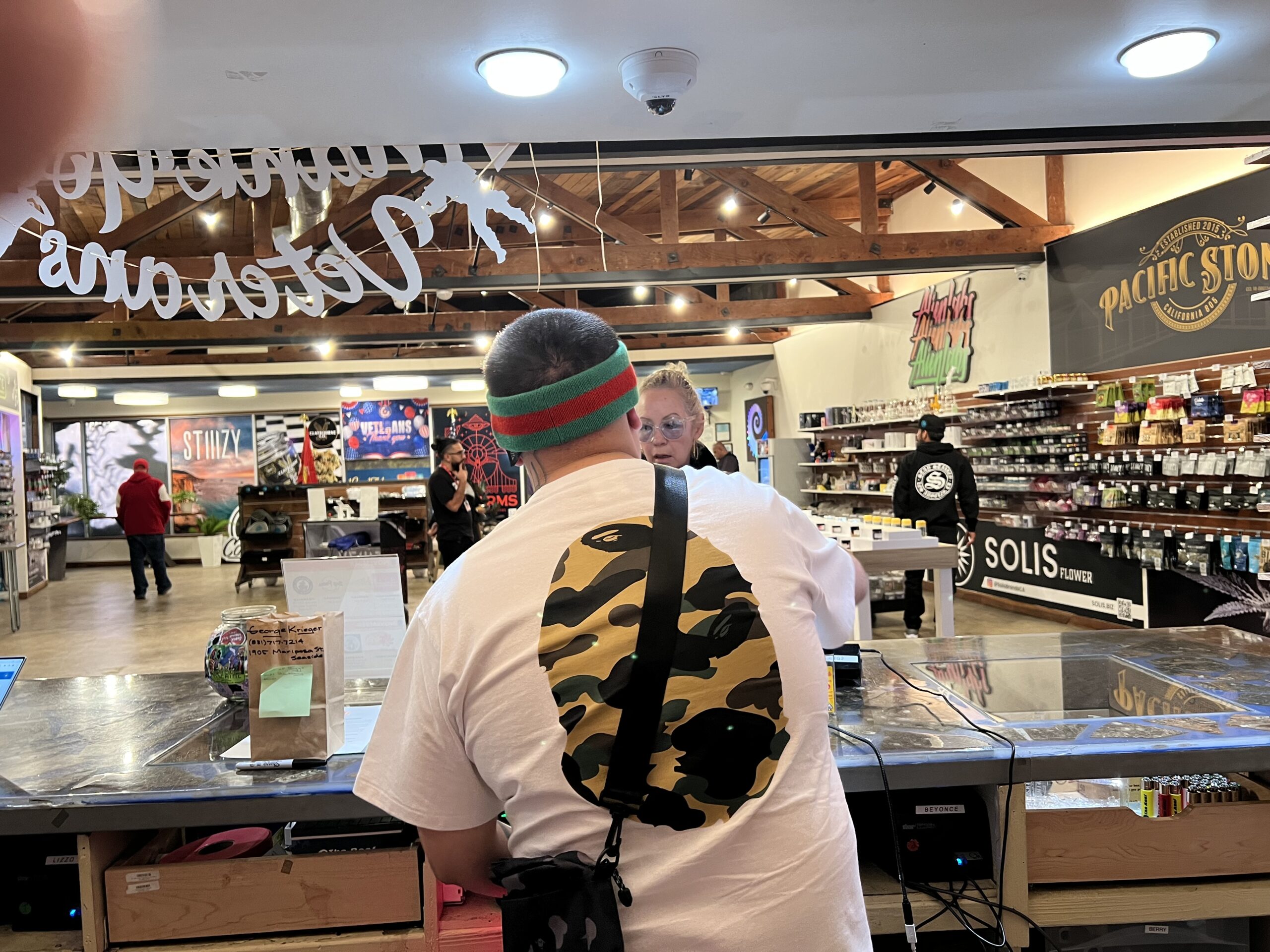 A man wearing a white t-shirt with a camouflage design and a green and red headband is interacting with a woman across a counter in a retail store